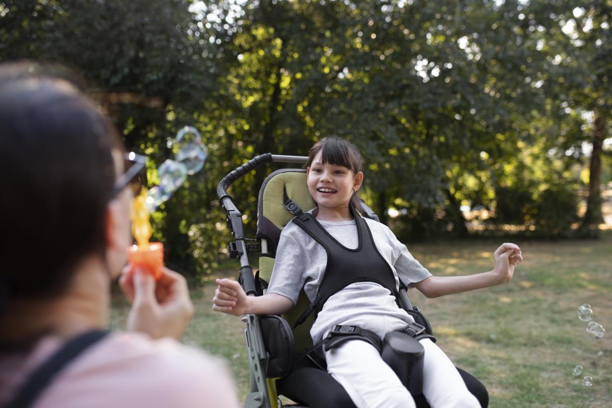Petite filles dans un fauteuil roulant poussette qui sourit devant une personne qui fait des bulles de savon